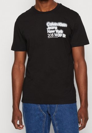 T-shirt en coton noir avec texte graphique blanc : "Calvin Klein Jeans New York 205 W39 St." Manches courtes et col rond.