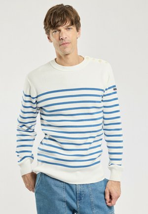 Homme portant un pull blanc à rayures horizontales bleues et un jean bleu clair, debout devant un fond uni clair.