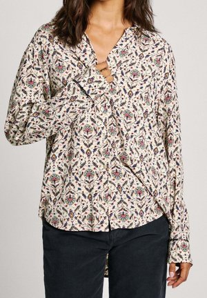 Vrouw draagt een crèmekleurige blouse met lange mouwen en veelkleurige bloemmotieven en zwarte broek, houdt de blouse bij de borst vast.