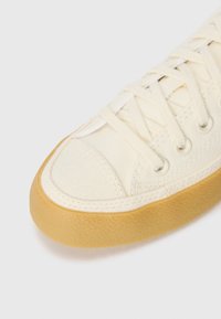 Canvas sneaker in off-white with a textured rubber sole. Features white laces and eyelets, rounded toe, and stitching details.