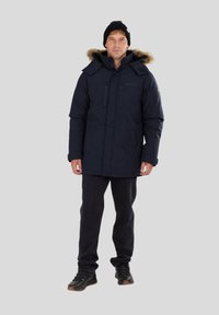 Parka bleu marine avec capuche en fausse fourrure, deux poches poitrine et rabat tempête. Portée avec un pantalon noir et des bottes de randonnée noires.