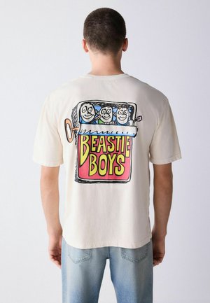 Rückansicht einer Person, die ein weißes T-Shirt mit einer bunten Cartoon-Aufziehbox trägt, die drei lächelnde Gesichter hält, und dem Text "BEASTIE BOYS".