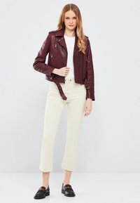 Bordeaux leren motorjack met zilveren ritsen, slim fit ontwerp en cropped lengte, gecombineerd met crèmekleurige rechte broek en zwarte instappers.