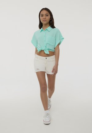 Camicia verde menta a maniche corte con bottoni e nodo frontale, abbinata a pantaloni shorts in denim bianco sbiadito e sneakers bianche.