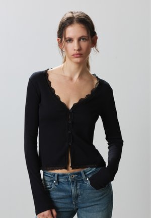 Jeune femme aux cheveux bruns attachés portant une chemise noire à manches longues boutonnée avec un bord en dentelle et un jean bleu, debout devant un fond uni.