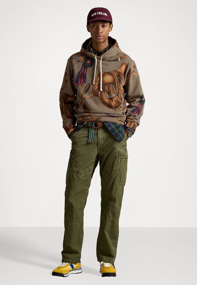 Jovem com um moletom estampado, equipado com acessórios de cavalo, calças cargo verde-oliva, sapatilhas amarelas, uma camisa de xadrez e um boné bordeaux com "New England".