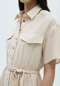 Abito camicia beige con scollatura a colletto, maniche corte, due tasche sul petto, bottoni in tono oro e cintura regolabile.