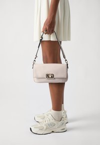 Borsa a mano in pelle rosa chiaro, dalla forma rettangolare, con hardware in argento e una tracolla removibile, tenuta in mano accanto a un paio di sneakers bianche.