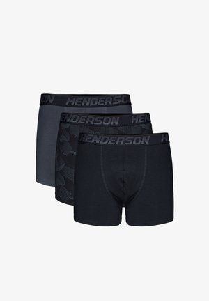 Oberkörperfreier Mann mit kurzen braunen Haaren, der schwarze Boxershorts mit gebrandetem Bund trägt, steht vor einem einfarbigen hellen Hintergrund.