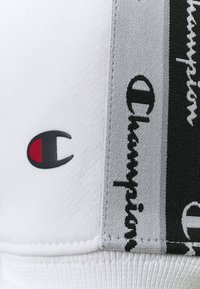 Weißer Stoff mit schwarzen, grauen und weißen vertikalen Streifen, der mit schwarzem "Champion"-Text und einem kleinen schwarzen und roten Champion-Logo-Patch verziert ist.