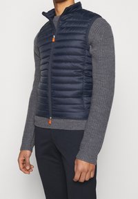 Navy puffer vest with quilted texture, high collar, front zipper, and orange zipper pull, worn over a gray knit sweater.
