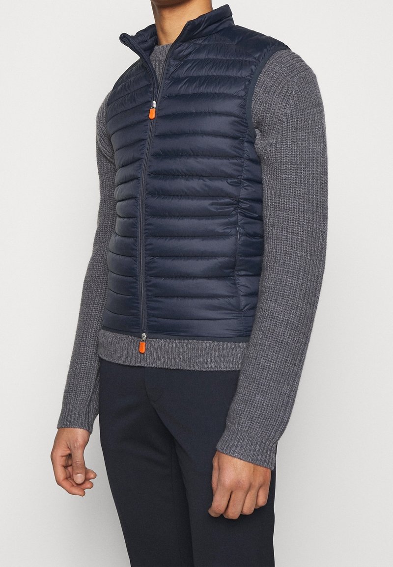 Navy puffer vest with quilted texture, high collar, front zipper, and orange zipper pull, worn over a gray knit sweater.