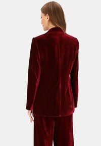 Burgundowa aksamitna marynarka o dopasowanym kroju, z gładką fakturą, usztywnionymi ramionami i pojedynczym rozcięciem na plecach.