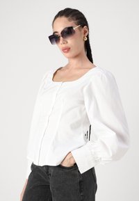 Biely blúzka s puff-sleeve rukávmi, štvorcovým výstrihom, predným gombíkom a nahrnutými manžetami, v kombinácii s oversized tmavými slnečnými okuliarmi a čiernymi džínsami.