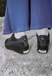 Černé semišové platformové tenisky s texturovanými detaily, na patě s logem PUMA a bílou gumovou podrážkou, doplněné o zkrácené modré kalhoty.