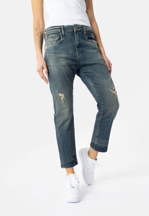 Kvinde iført falmede, slidte blå jeans og hvide sneakers, stående med det ene ben let bøjet mod en ensfarvet baggrund.