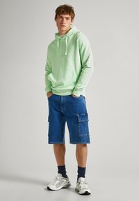 Sudadera verde claro con bolsillo frontal, combinada con pantalones cortos cargo de mezclilla azules hasta la rodilla que tienen bolsillos laterales. Zapatillas blancas y calcetines azul marino.