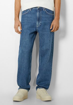 Man draagt rechte blauwe spijkerbroek en licht crèmekleurige sneakers, staand tegen een effen witte achtergrond.