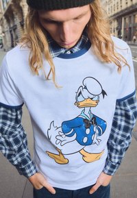 Bijela majica s plavom i crnom ilustracijom Donalda Ducka koji nosi plavi kaput i leptir mašnu. Dugi košuljski rukavi na kockice vidljivi ispod.