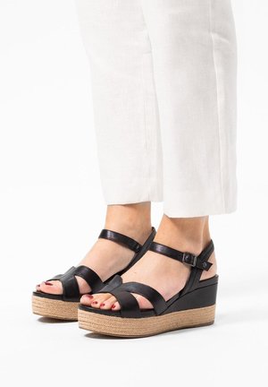 Black leather wedge sandals with a woven jute platform, featuring crossed straps and ankle buckle. Smooth texture and open toe design.
