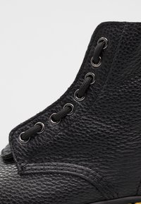 Gros plan d'une botte en cuir noir texturé avec des lacets noirs passés dans des œillets métalliques sur un fond clair.