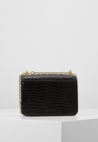 Svart läderhandväska med krokodilstruktur och guldkedjesnodd som vilar på en vit yta mot en grå bakgrund.