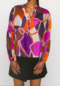 Blusa de manga larga colorida con grandes patrones florales en púrpura, naranja y beige, con un escote abotonado y puños fruncidos.