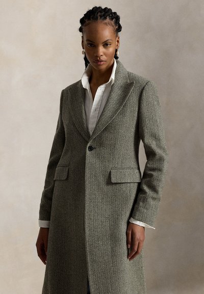 Femme aux cheveux tressés, portant des boucles d'oreilles en or et un long manteau gris à chevrons par-dessus une chemise blanche boutonnée, debout devant un fond uni.