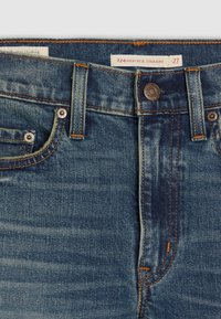 Jean droit taille haute en denim bleu avec coutures orange, bouton visible, passants de ceinture et étiquette de taille indiquant 27.