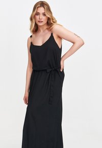 Kobieta w czarnej sukience na cienkich ramiączkach z kokardą w talii, stojąca pewnie z jedną ręką na biodrze i delikatnym uśmiechem.