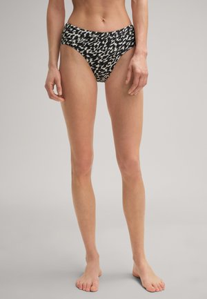 Eine weibliche Model trägt schwarze und weiße gemusterte hoch geschnittene Bikiniunterteile und steht barfuß vor einem schlichten grauen Hintergrund.