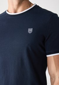 Tamno plava majica s kratkim rukavima, okruglim bijelim ovratnikom i bijelim detaljima na manžetama. Ima mali izvezeni logotip na prsima.