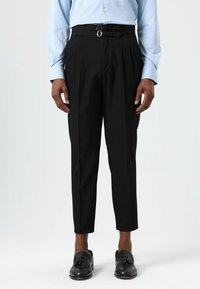 Pantalon noir taille haute avec plis et double boucle en accent. Porté avec une chemise bleu clair à manches longues et des mocassins noirs.