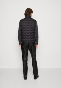 Czarna pikowana kamizelka puchowa z wysokim kołnierzem, w połączeniu z szarą koszulą z długim rękawem i czarnymi skórzanymi spodniami, widziana od tyłu.