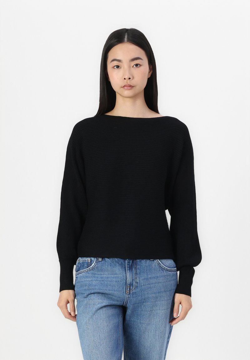 Zwarte gebreide trui met een bootnek, ontspannen pasvorm en lange mouwen, gecombineerd met lichtblauwe denim jeans. Gladde textuur, geen patronen.