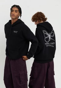 Le felpe nere presentano una tasca frontale e testo bianco sul petto, con una grafica di una farfalla sul retro, abbinate a pantaloni cargo viola.