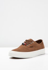 Baskets en daim marron avec semelle en caoutchouc blanche, dotées d'œillets métalliques et de lacets décoratifs bleus et rouges, design minimaliste.