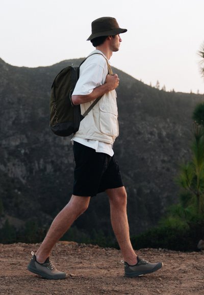 Conjunto de senderismo que presenta un chaleco beige sobre una camisa blanca, pantalones cortos negros, una mochila oliva y zapatillas deportivas grises, con un fondo montañoso.
