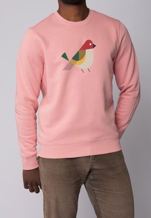 Uomo con felpa rosa con disegno geometrico di un uccello, abbinata a pantaloni di velluto a coste marroni, in piedi su uno sfondo grigio chiaro.