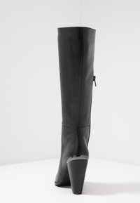 Bota de cuero negro hasta la rodilla con un acabado texturizado, forma cónica y tacón cónico. Cuenta con un cierre lateral para facilitar su uso.