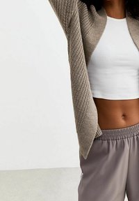 Beige pletený cardigan s diagonálně žebrovanou texturou, otevřený vpředu, nošený přes bílý zkrácený top, doplněný o světle šedé kalhoty s elastickým pasem.