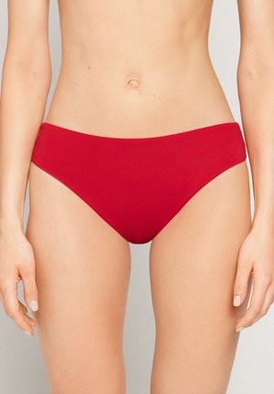 Kobieta w czerwonych, gładkich majtkach bikini, ukazująca odsłonięty brzuch i górną część ud, z rękami swobodnie opuszczonymi wzdłuż ciała na neutralnym tle.