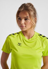Neon grøn atletisk t-shirt med sorte skulderakcenter og logo. Glat tekstur, korte ærmer, rund halsudskæring, designet til aktiv brug.