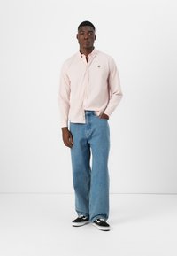 Light pink long-sleeve shirt with button-down collar and chest logo, paired with wide-leg blue denim jeans and black sneakers.