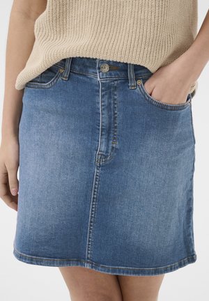 Kvinde iført lys blå denim nederdel med forreste knap og lynlås, hånd i højre lomme, kombineret med beige striktop.