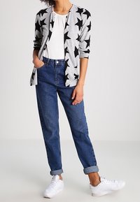 Cardigan grigio con motivo a stelle nere, top bianco e jeans blu con risvolto. Indossato con sneakers bianche, creando un outfit casual a strati.