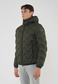 Giubbotto trapuntato verde con cappuccio, dotato di un patch logo in gomma e tasche con zip. Visibili polsini e orlo a coste.