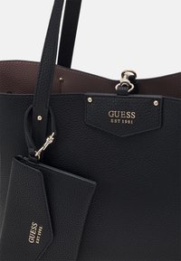 Zwarte gestructureerde leren tote bag met gouden hardware en een label met "GUESS EST 1981." Inclusief een afneembare kaarthouder.