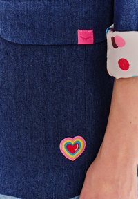 Denimowa bluzka z granatową teksturą, z różową naszywką i wielokolorową łatą w kształcie serca po lewej stronie blisko kieszeni.