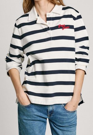 Kobieta ubrana w biało-czarną poziomo w pasy bluzkę polo z długim rękawem z czerwonym logo "Pepe Jeans" i niebieskie dżinsy, ręce w kieszeniach.